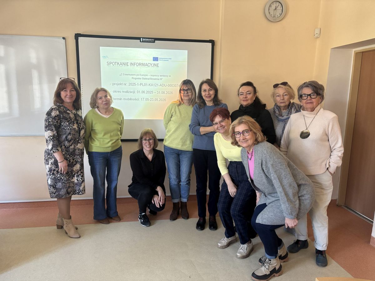Na zdjęciu widać grupę dziesięciu kobiet znajdujących się w sali, stojących i kucających przed tablicą multimedialną z wyświetloną prezentacją. Na slajdzie widnieje tytuł „Spotkanie informacyjne” oraz informacje dotyczące projektu Erasmus+ i planowanej mobilności.