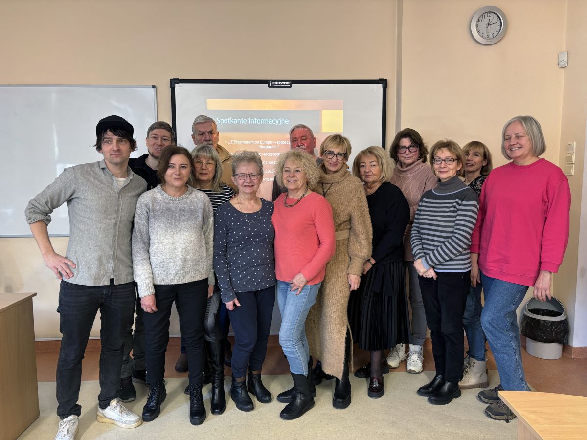 Na zdjęciu widać grupę czternastu dorosłych osób stojących w sali przypominającej klasę lub salę szkoleniową. Uśmiechają się do zdjęcia i są ubrani w codzienne, jesienno-zimowe stroje w stonowanych kolorach. Za nimi znajduje się ekran z napisem „Spotkanie informacyjne”, obok biała tablica i zegar na ścianie.